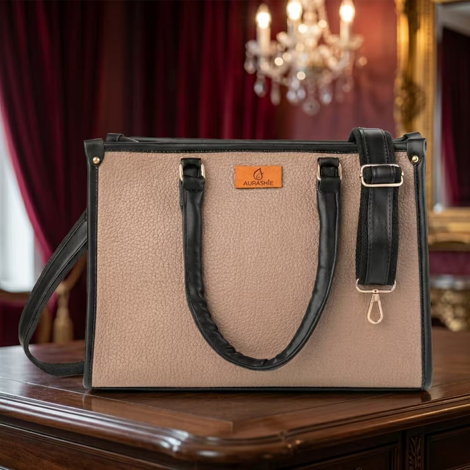 Beige handbag with black handles and strap on a wooden surface with a chandelier in the background