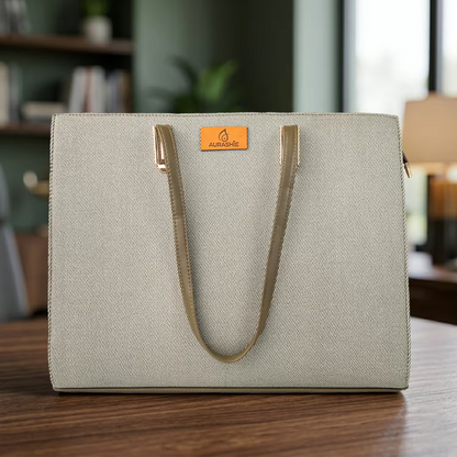 Light gray tote bag with gold handles on a wooden surface, blurred indoor background
